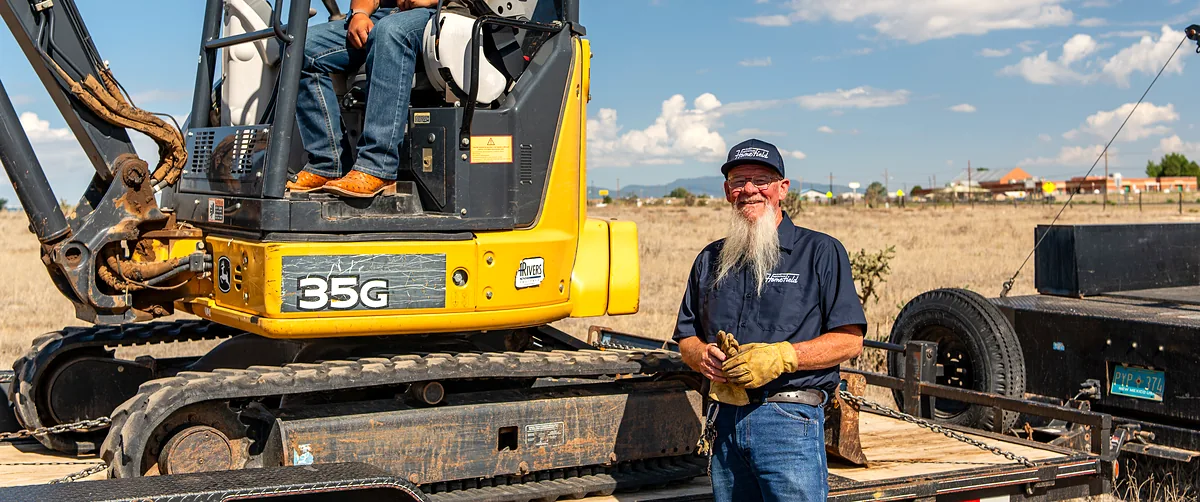 HomeField East Mountain operator in a compact excavator cab while a smiling crew member waits beside the trailer