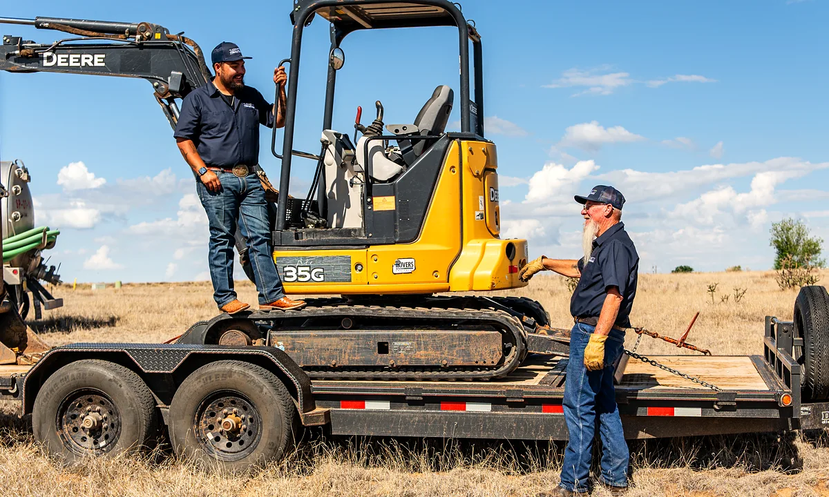 HomeField East Mountain crew member climbing onto a compact excavator on a flatbed trailer while a coworker secures the load
